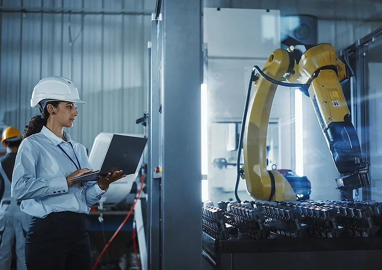 Industrial engineer at work. Hispanic woman using a laptop to monitor a robotic arm in a high-tech metal factory to ensure precise automation and efficiency in the production process