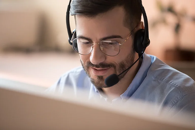 Telemarketer—telemarketing employee wearing a wireless headset, conducting a video conference with an online client via computer; male helpline operator working in a customer service office