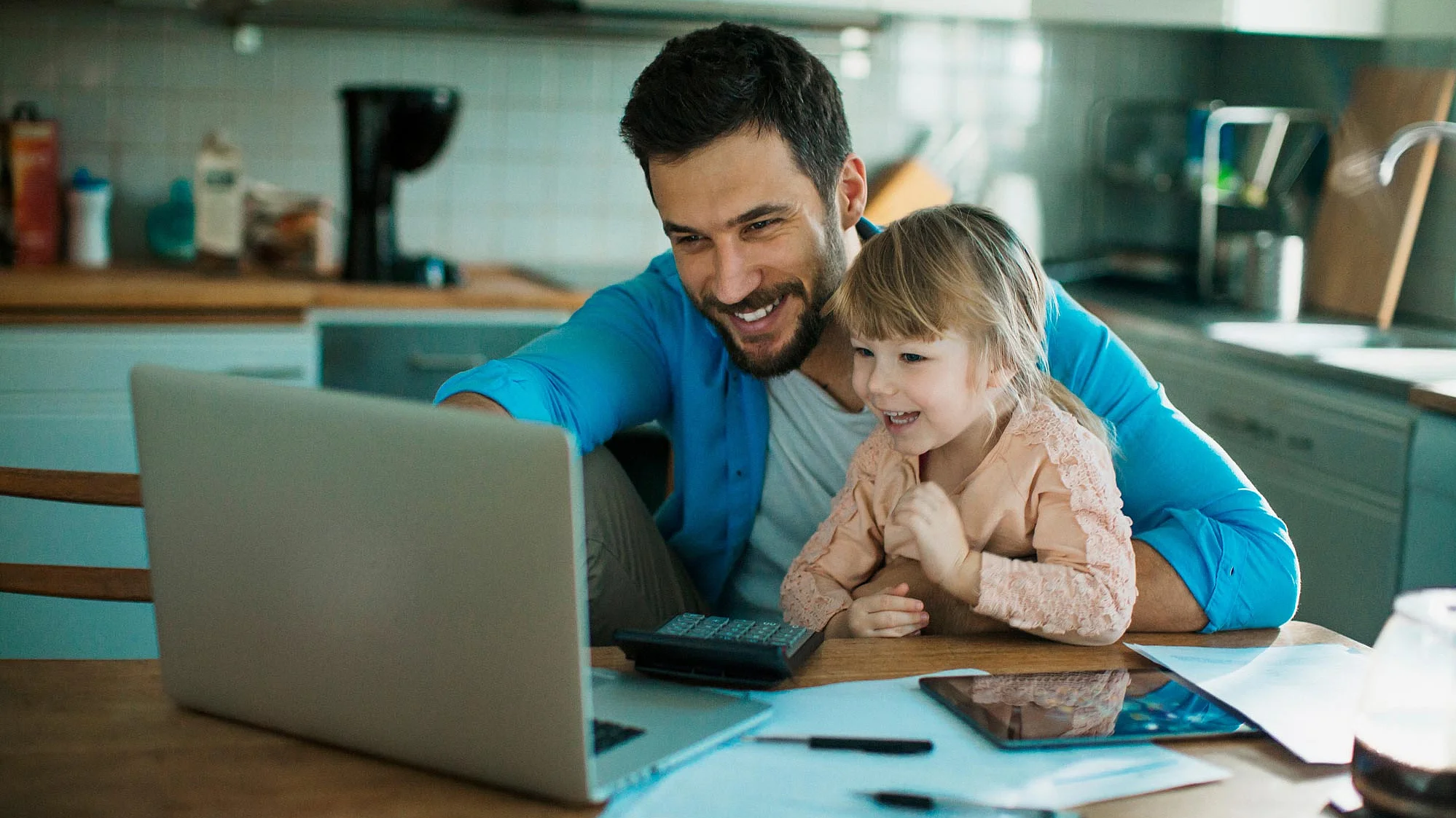 A smiling young man sitting at his desk with his young daughter, both looking at the laptop
