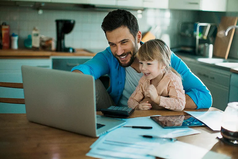 A smiling young man sitting at his desk with his young daughter, both looking at the laptop